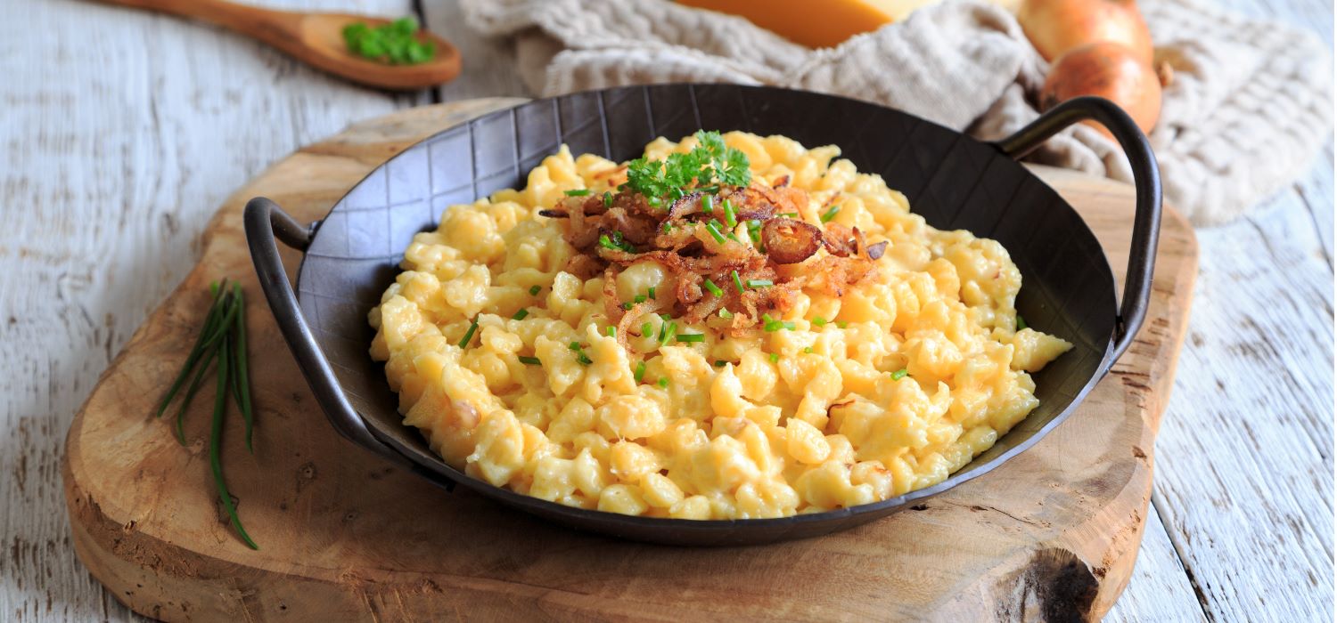 Käsespätzle mit Röstzwiebeln garniert mit Kräutern serviert in einer dunklen topfähnlichen Schüssel auf einem Holzbrett.