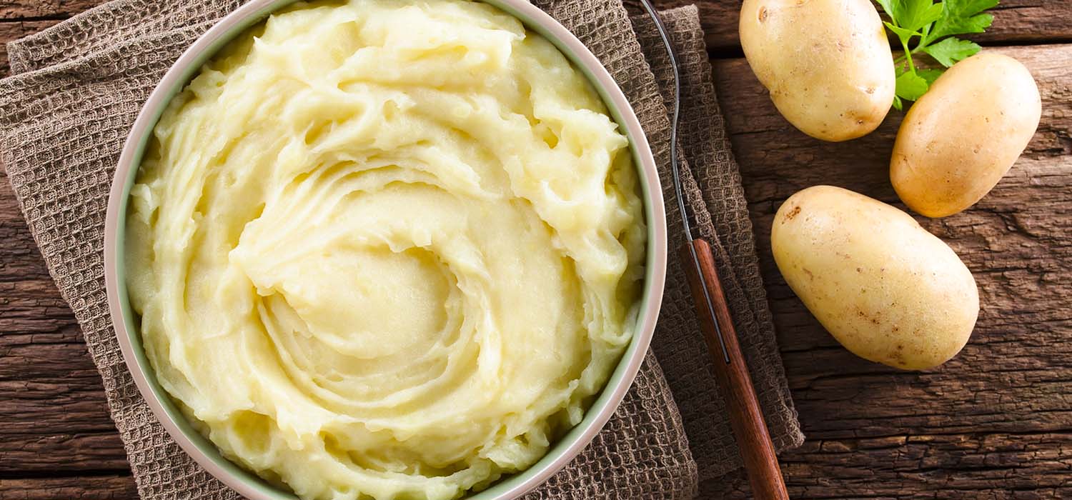 Schüssel Kartoffelpüree mit einer Gabel und danebenliegenden Kartoffeln.