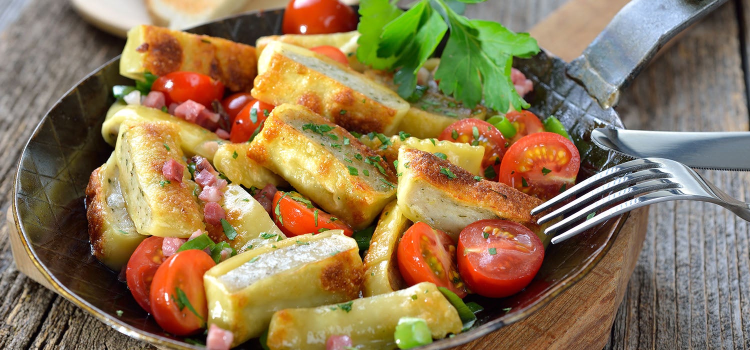 Maultaschen angerichtet in einer Pfanne auf einem Holzbrett mit Tomaten, Speck und Kräutern garniert mit Frühlingszwiebeln.