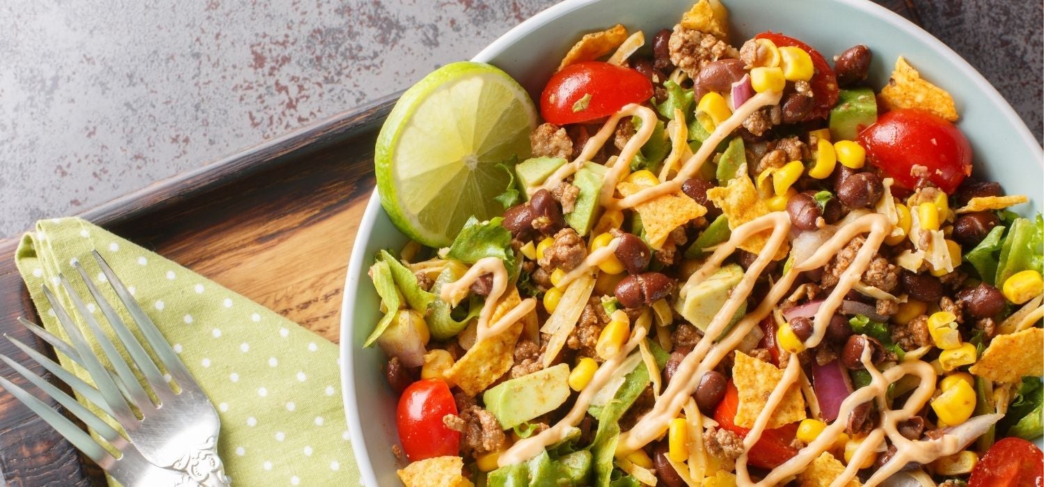 Taco-Salat mit Hackfleisch, Bohnen, Eisbergsalat, Tomaten, Zwiebeln, Käse und Avocado garniert mit einer Limette serviert in einer Schüssel auf einem Tablett.